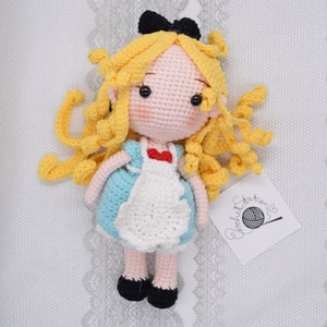 May include: A crocheted doll of Alice from Alice in Wonderland. She has long blonde hair, a blue dress with a white apron, and black shoes. She is wearing a red bow on her chest.