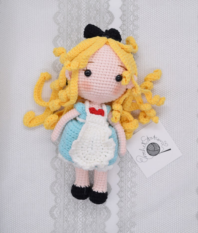 May include: A crocheted doll of Alice from Alice in Wonderland. She has long blonde hair, a blue dress with a white apron, and black shoes. She is wearing a red bow on her chest.