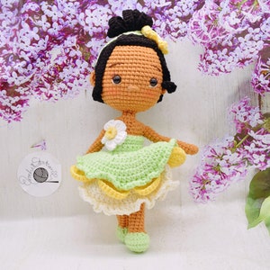 May include: A crocheted doll with brown skin, black hair, and a green dress with yellow accents. The doll is wearing a flower on her dress and has a sweet expression on her face.
