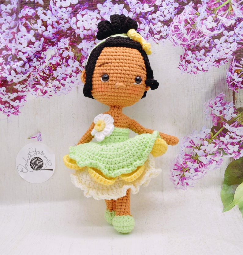 May include: A crocheted doll with brown skin, black hair, and a green dress with yellow accents. The doll is wearing a flower on her dress and has a sweet expression on her face.