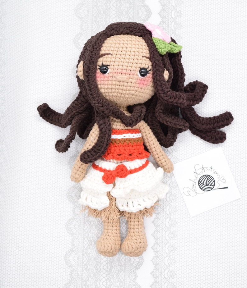 May include: Crocheted doll with brown hair, a red and white top, and a white skirt. The doll is wearing a flower in her hair and has a light brown complexion.