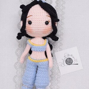 May include: Crocheted doll with black hair, a blue outfit, and a yellow belt. The doll has a pink face with black eyes and a small smile. The doll is sitting on a white surface with a white lace pattern.