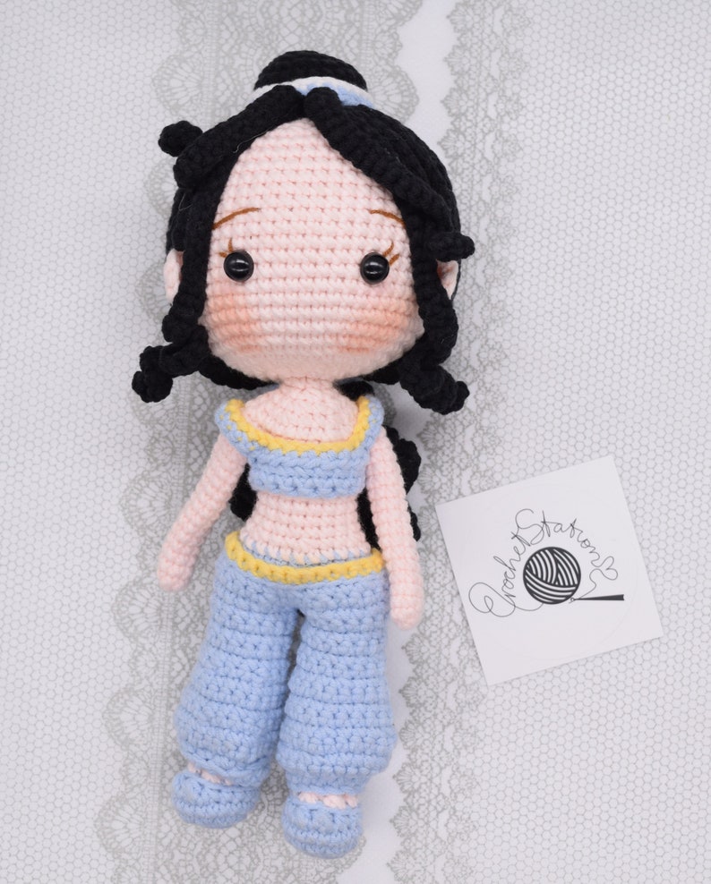 May include: Crocheted doll with black hair, a blue outfit, and a yellow belt. The doll has a pink face with black eyes and a small smile. The doll is sitting on a white surface with a white lace pattern.