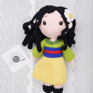 May include: A crocheted doll with black hair, a yellow dress, green and blue accents, and a white flower in her hair.