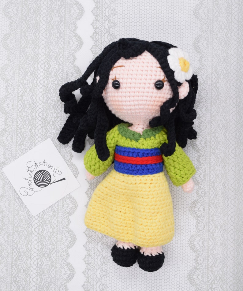 May include: A crocheted doll with black hair, a yellow dress, green and blue accents, and a white flower in her hair.