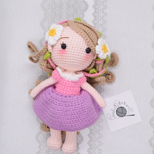 May include: A crocheted doll with brown hair, a pink dress, and a purple skirt. The doll is wearing a flower crown with white flowers and green leaves. The doll is sitting on a white background with a crocheted label that says "crochet stories".