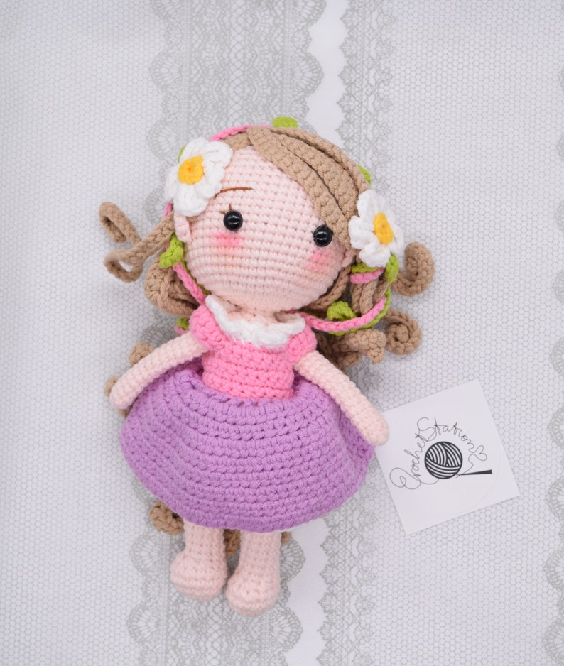 May include: A crocheted doll with brown hair, a pink dress, and a purple skirt. The doll is wearing a flower crown with white flowers and green leaves. The doll is sitting on a white background with a crocheted label that says "crochet stories".