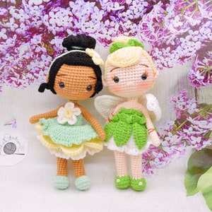 May include: Two crocheted dolls, one with brown skin and a green dress, the other with fair skin and a green dress with wings. Both dolls have brown eyes and black hair.