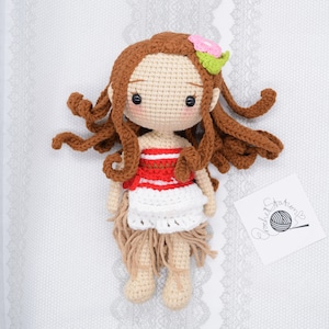 May include: Crocheted doll with brown hair, a red and white striped top, a white skirt, and a flower in her hair. The doll is lying on a white background with a crocheted pattern.