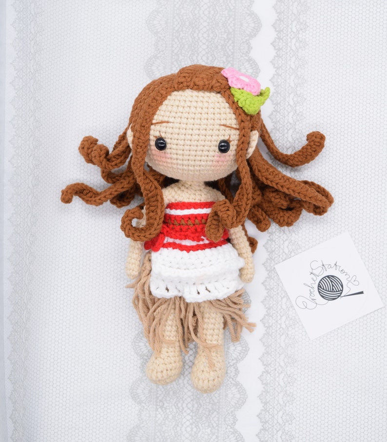May include: Crocheted doll with brown hair, a red and white striped top, a white skirt, and a flower in her hair. The doll is lying on a white background with a crocheted pattern.