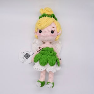 May include: A crocheted fairy doll with yellow hair, green wings, and a green dress. The doll is standing on a white background.