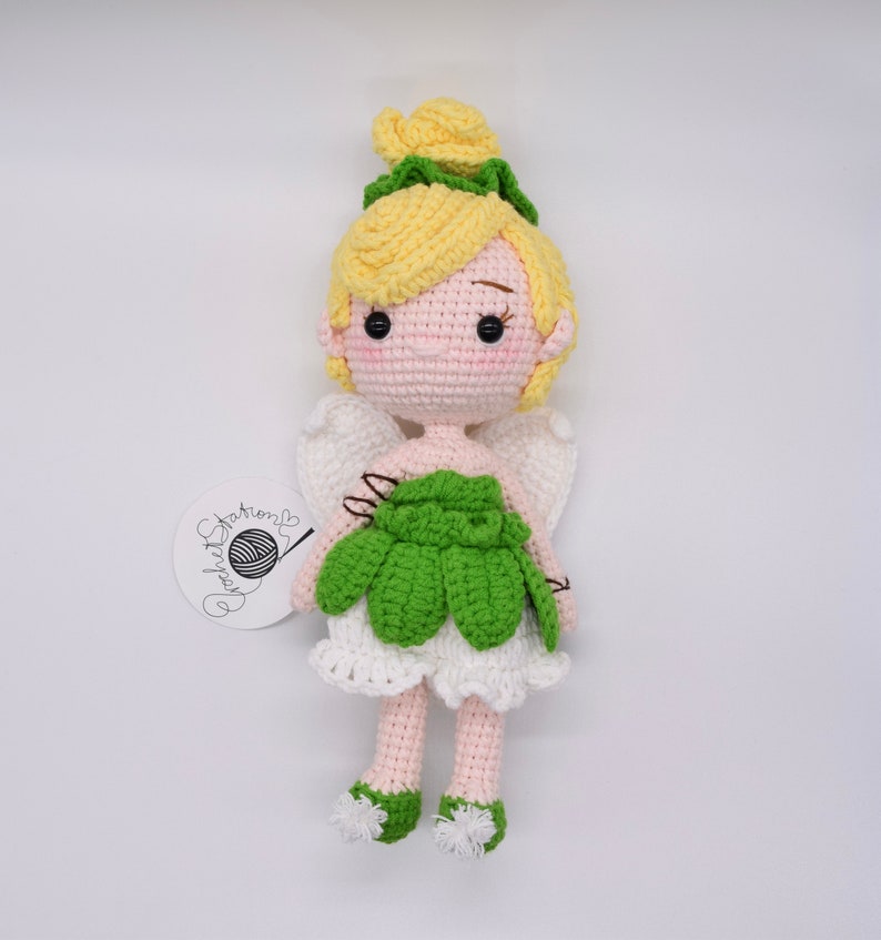 May include: A crocheted fairy doll with yellow hair, green wings, and a green dress. The doll is standing on a white background.