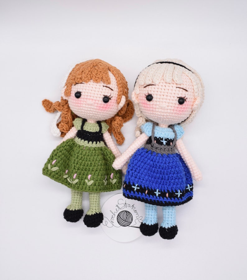 May include: Two crocheted dolls, one with brown hair and a green dress, the other with blonde hair and a blue dress. Both dolls have black eyes and are wearing black shoes.