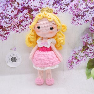 May include: A crocheted doll with blonde hair, a pink dress, and a crown. The doll is standing on a white surface with purple flowers in the background.