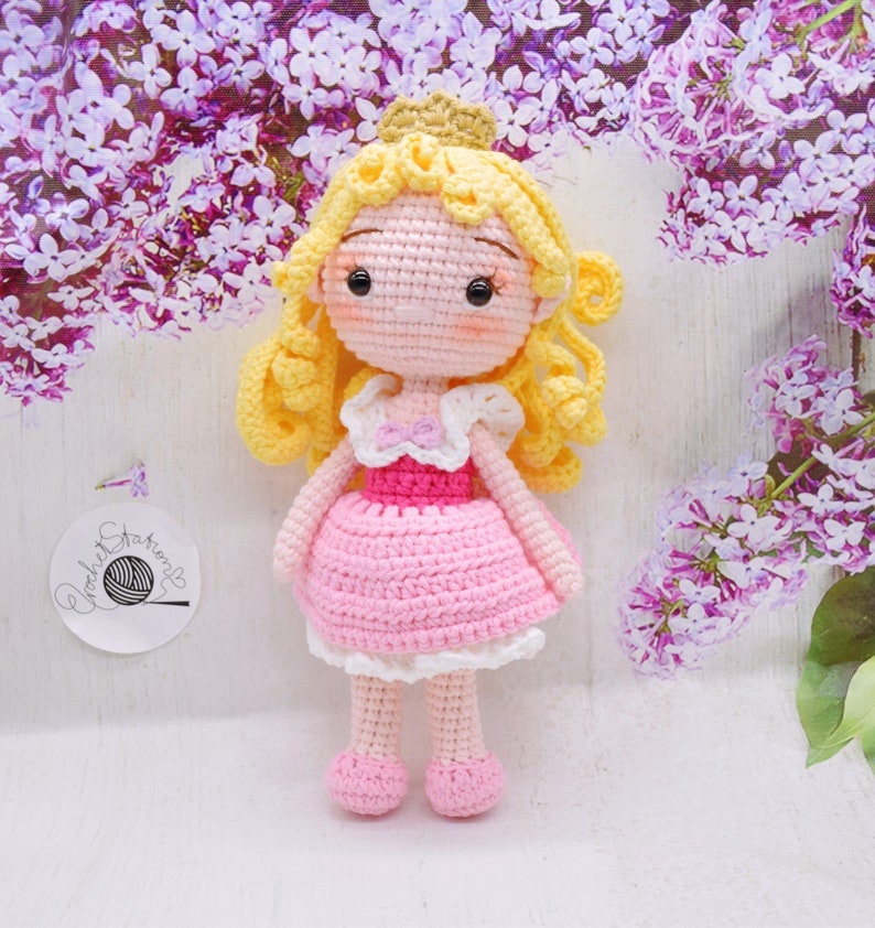 May include: A crocheted doll with blonde hair, a pink dress, and a crown. The doll is standing on a white surface with purple flowers in the background.