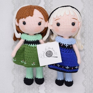 May include: Two crocheted dolls, one with brown hair and a green dress, the other with blonde hair and a blue dress. The dolls are standing on a white background with a crocheted pattern.