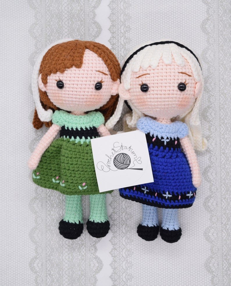 May include: Two crocheted dolls, one with brown hair and a green dress, the other with blonde hair and a blue dress. The dolls are standing on a white background with a crocheted pattern.