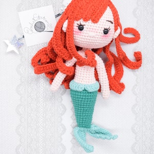 May include: A crocheted mermaid doll with long red hair, a pink torso, and a teal tail. The doll is lying on a white lace background.