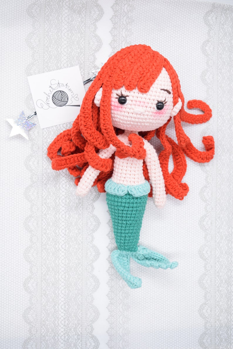May include: A crocheted mermaid doll with long red hair, a pink torso, and a teal tail. The doll is lying on a white lace background.