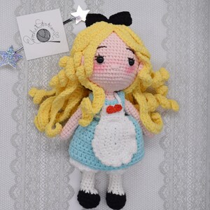 May include: A crocheted doll of Alice from Alice in Wonderland. She has long blonde hair, a blue dress with a white apron, and black shoes. She is wearing a black bow on her head.