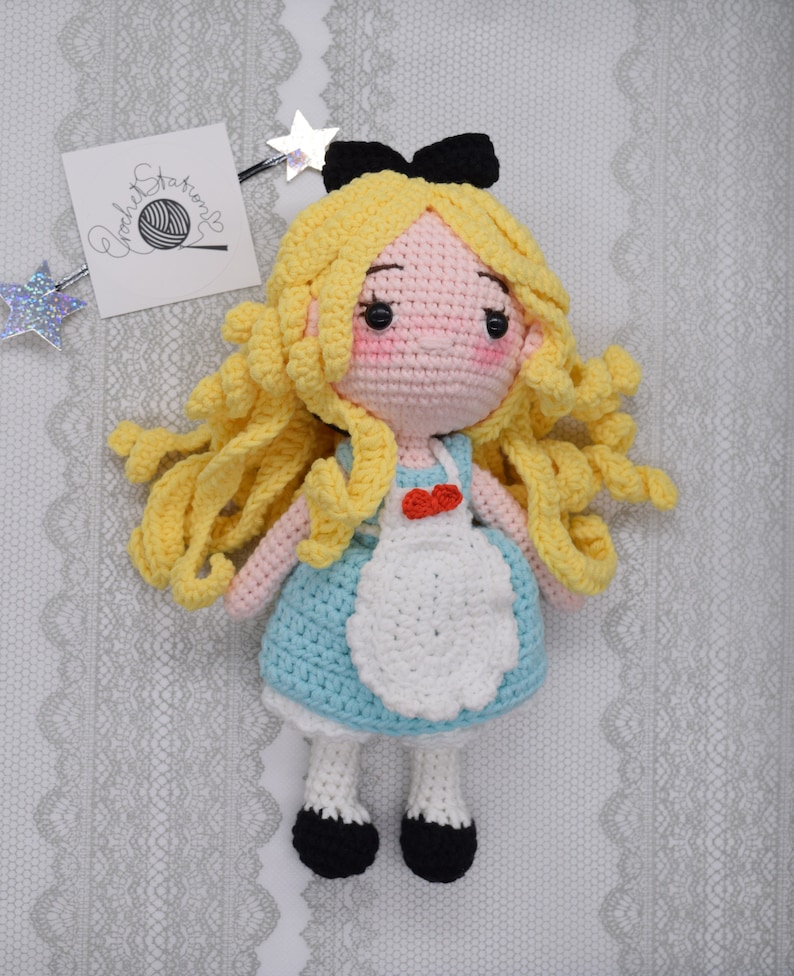 May include: A crocheted doll of Alice from Alice in Wonderland. She has long blonde hair, a blue dress with a white apron, and black shoes. She is wearing a black bow on her head.