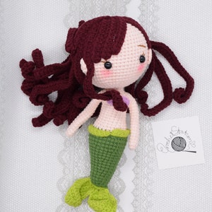 May include: A crocheted mermaid doll with long, curly burgundy hair, a pink torso, a green tail, and a purple seashell bra. The doll is lying on a white lace background.