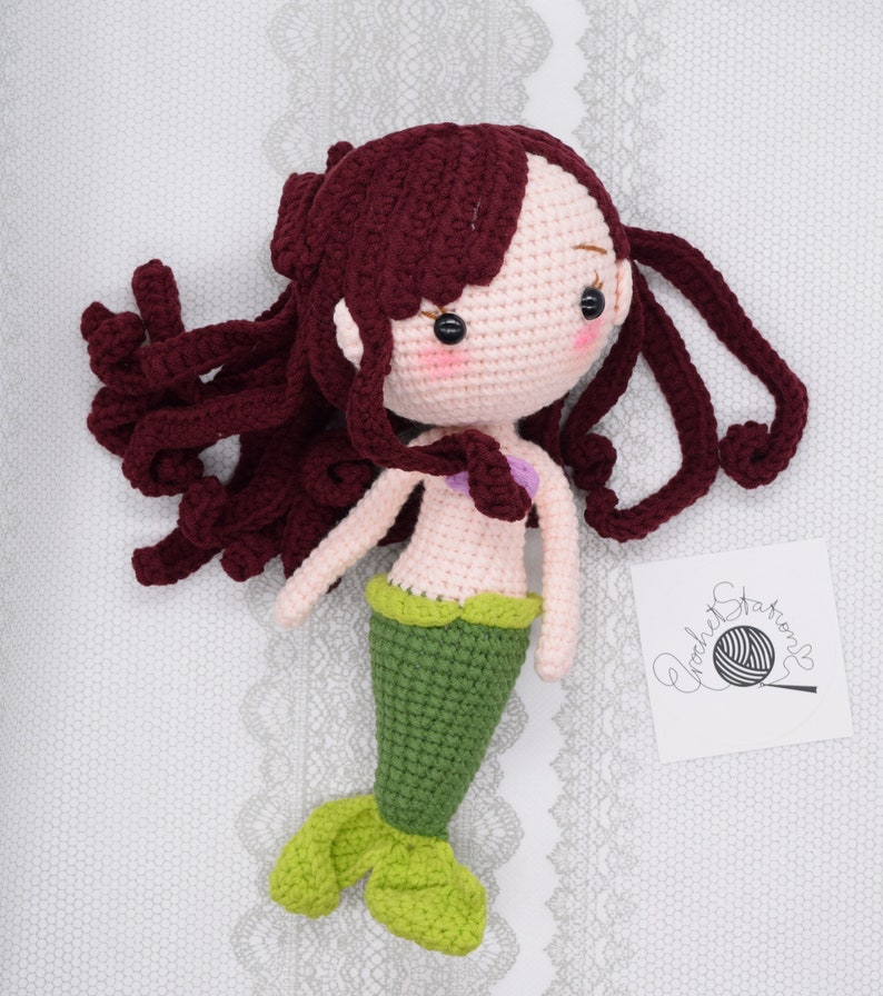 May include: A crocheted mermaid doll with long, curly burgundy hair, a pink torso, a green tail, and a purple seashell bra. The doll is lying on a white lace background.