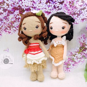 May include: Two crocheted dolls, one with brown hair and a red and white dress, the other with black hair and a brown dress. Both dolls have brown eyes and are standing on a white surface.