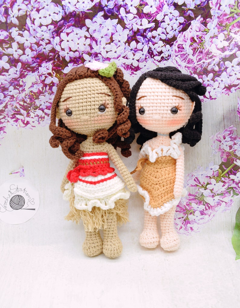 May include: Two crocheted dolls, one with brown hair and a red and white dress, the other with black hair and a brown dress. Both dolls have brown eyes and are standing on a white surface.