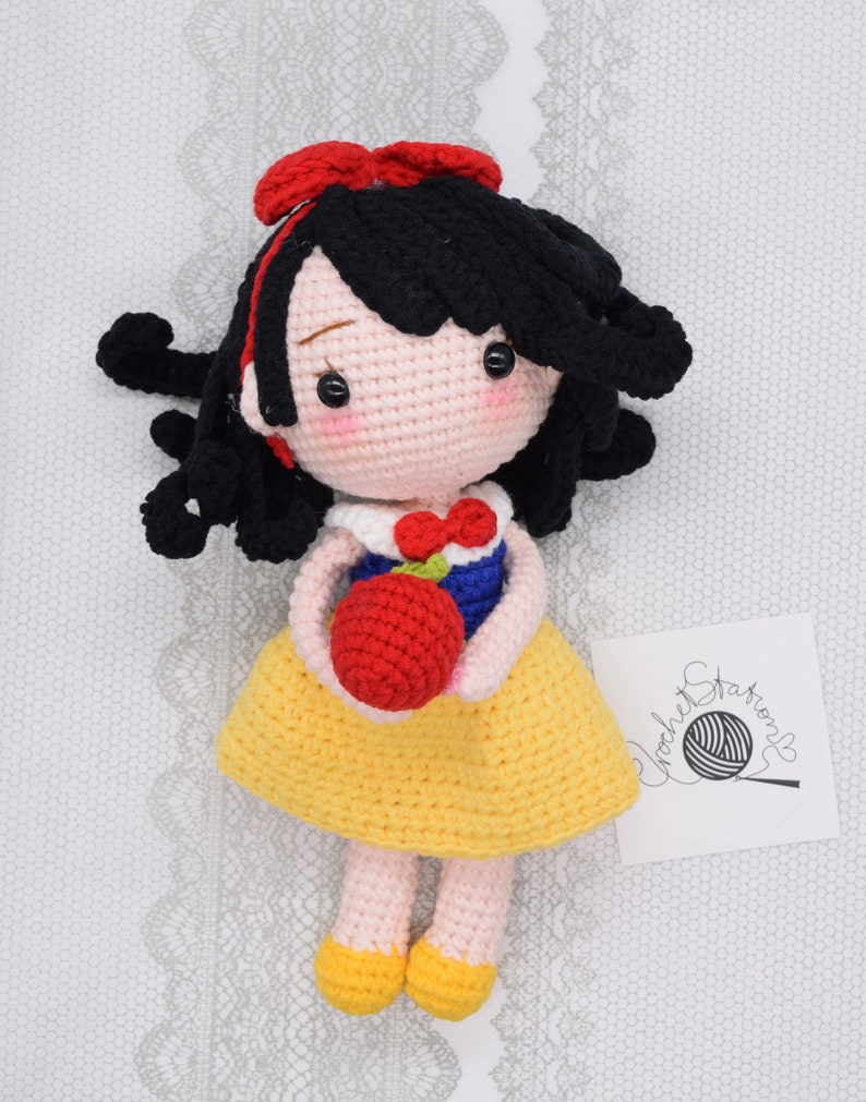 May include: A crocheted Snow White doll with black hair, a red bow, a yellow dress, and a red apple. The doll is holding the apple in her hands. The doll is sitting on a white background with a lace pattern.