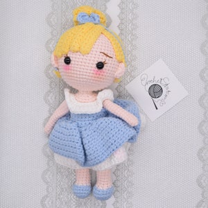 May include: A crocheted doll with blonde hair, a blue dress, and blue shoes. The doll is wearing a blue bow in her hair. The doll is sitting on a white background with a crocheted pattern.