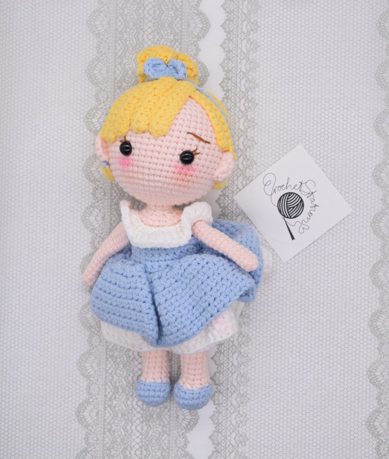 May include: A crocheted doll with blonde hair, a blue dress, and blue shoes. The doll is wearing a blue bow in her hair. The doll is sitting on a white background with a crocheted pattern.