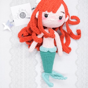May include: A crocheted mermaid doll with long red hair, a pink torso, and a teal tail. The doll is lying on a white lace background.