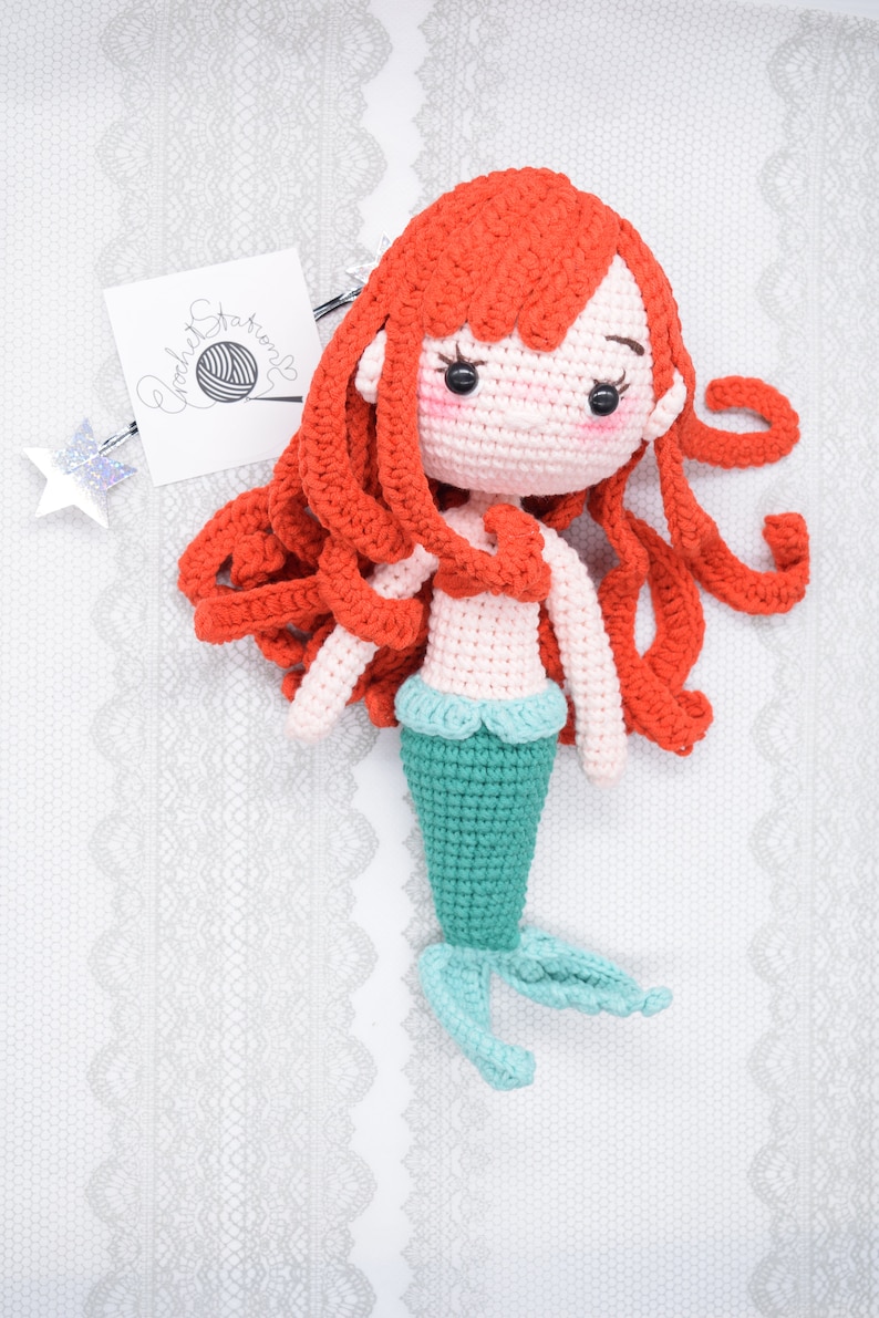 May include: A crocheted mermaid doll with long red hair, a pink torso, and a teal tail. The doll is lying on a white lace background.