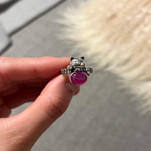 May include: A silver-toned ring featuring a cat design holding a large, oval, magenta-colored stone. The cat's face is detailed, and the ring band has decorative elements. The ring is held up against a blurred background.