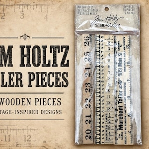 May include: A clear plastic package containing five wooden ruler pieces with various printed designs. The pieces feature black markings, numbers, and text including 'Merchant Tailor' and '209 Main St.' The packaging is labeled 'Tim Holtz idea-ology'.
