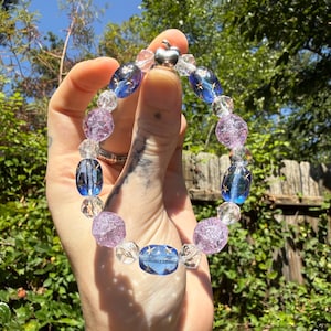 May include: A beaded bracelet featuring alternating translucent purple crackle glass beads, clear faceted crystal beads, and blue oval beads decorated with small gold star patterns, finished with a small silver-toned apple-shaped charm.