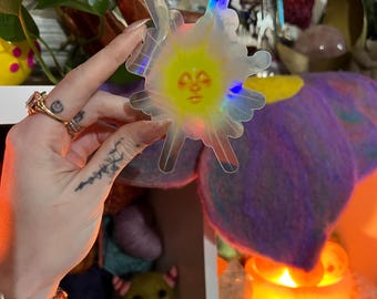 Puede incluir: Una pegatina transparente con forma de sol con una cara amarilla y bordes radiantes. La pegatina tiene un efecto holográfico, que refleja múltiples colores. La imagen es de una pegatina, posiblemente para un diario o portátil, con un diseño caprichoso.