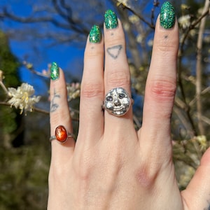 May include: A silver ring with a face-shaped stone set in the center. The ring is on a person's finger, and the background is a blurry image of green leaves and white flowers.