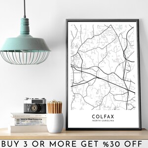 May include: Black and white line art map print of Colfax, North Carolina in a white frame on a wooden shelf. The text "BUY 3 OR MORE GET %30 OFF" is printed on the shelf.