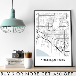 May include: A black and white map print of American Fork, Utah, in a black frame. The map shows the city's streets and roads. The text "AMERICAN FORK" and "UTAH" is printed below the map. The print is displayed on a shelf with a light fixture, a camera, and books.