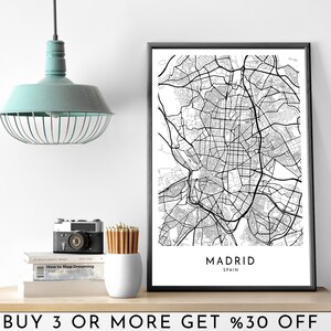 Op de afbeelding: Zwart-witte stadskaartprint van Madrid, Spanje in een zwart frame. De print staat op een plank met een lamp, een camera en boeken.