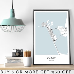 May include: A black and white city map print of Cadiz, Spain with a light blue background. The print is framed in a black frame and is hanging on a white wall. The text "Cádiz" and "España" is printed in black at the bottom of the map.