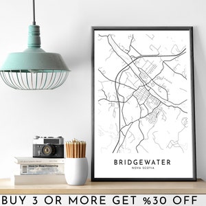 May include: Black and white line art map print of Bridgewater, Nova Scotia in a black frame. The text "BRIDGEWATER" and "NOVA SCOTIA" is printed below the map. The print is on a shelf with a light blue pendant light, a camera, pencils, and books.