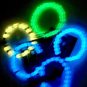 K&ouml;nnte beinhalten: Abstraktes Bild von leuchtenden LED-Lichtern in Gr&uuml;n, Gelb und Blau. Die Lichter sind in einem kreisf&ouml;rmigen Muster angeordnet und erzeugen eine dynamische und farbenfrohe Darstellung vor einem dunklen Hintergrund. Die Lichter scheinen durch d&uuml;nne Dr&auml;hte verbunden zu sein.