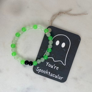 K&ouml;nnte beinhalten: Ein Armband mit gr&uuml;nen und durchsichtigen Perlen und zwei schwarzen Perlen. Ein schwarzes Etikett mit einer Geisterillustration und dem Text "You're Spooktacular" ist mit einer Schnur am Armband befestigt.