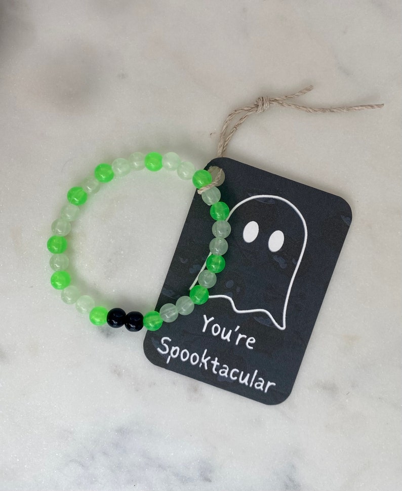 K&ouml;nnte beinhalten: Ein Armband mit gr&uuml;nen und durchsichtigen Perlen und zwei schwarzen Perlen. Ein schwarzes Etikett mit einer Geisterillustration und dem Text "You're Spooktacular" ist mit einer Schnur am Armband befestigt.