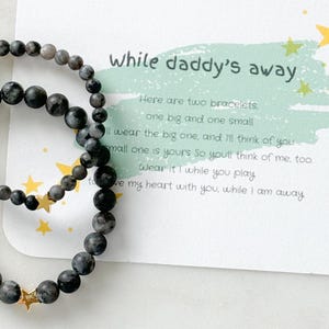 May include: Two black beaded bracelets with gold star charms and a card that reads "While daddy's away" with a poem about the bracelets.