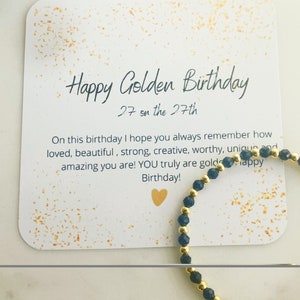 May include: A card with gold glitter and the text "Happy Golden Birthday 27 on the 27th" and a message about remembering how loved, beautiful, strong, creative, worthy, unique and amazing you are. The card is next to a black and gold beaded bracelet.