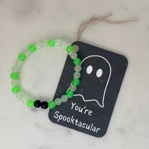 K&ouml;nnte beinhalten: Ein Armband mit abwechselnd gr&uuml;nen und durchsichtigen Perlen und zwei schwarzen Perlen. Ein schwarzes Etikett mit einer Geisterillustration und dem Schriftzug "You're Spooktacular" ist mit Bindfaden am Armband befestigt. Das Armband liegt auf einer wei&szlig;en Oberfl&auml;che.